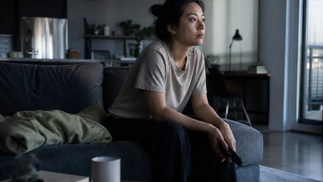 A person sits on a sofa in quiet daylight, phone nearby, looking depleted rather than distressed.